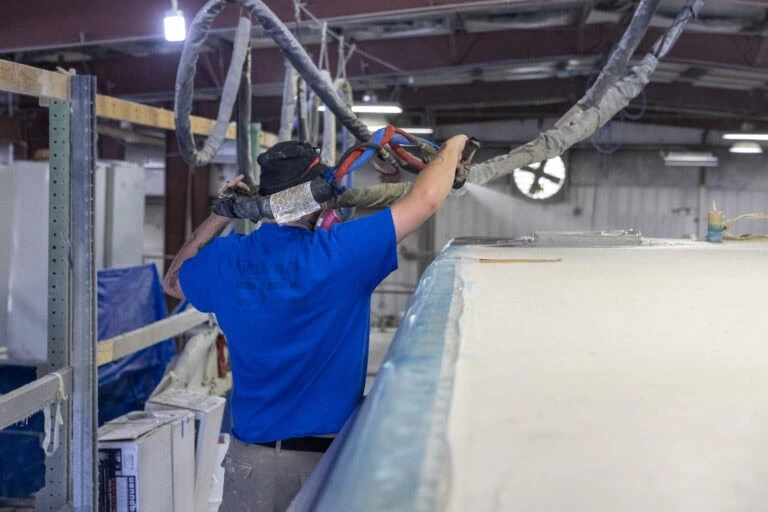 A technician wearing a blue shirt sprays a protective polyurea coating onto the roof of an RV inside a manufacturing facility, using a large spray rig connected to overhead hoses.