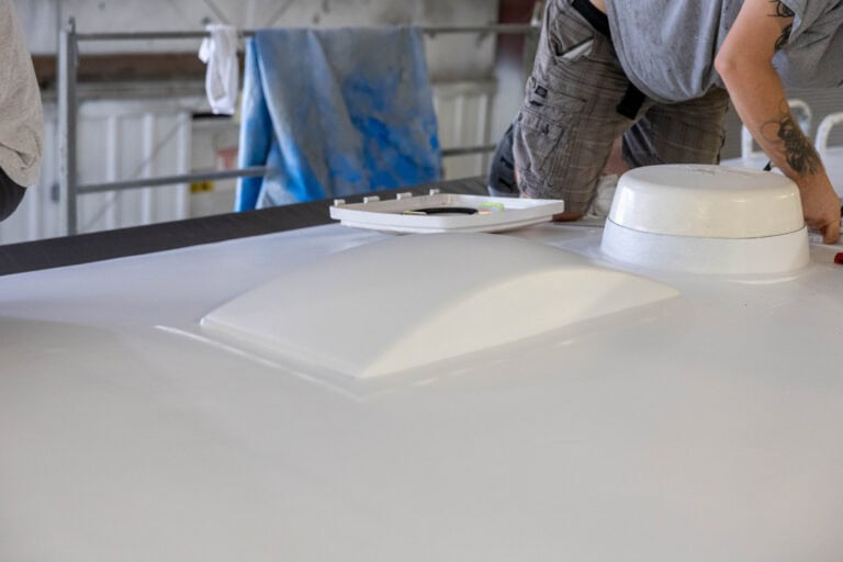 Technician working on the roof of an RV during a roof installation process, with a newly coated white roof surface, roof vent, and skylight visible in the foreground.