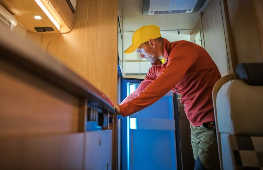 Man wearing a yellow cap and red long-sleeve shirt checks inside a refrigerator in an RV camper van.